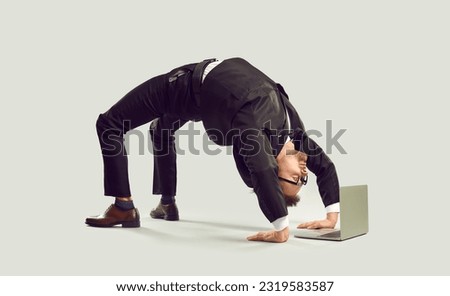 Extravagant curious businessman standing in bridge position looking at laptop screen on gray background. Crazy young man in suit stands on his hands and feet in yoga pose bridge near laptop.