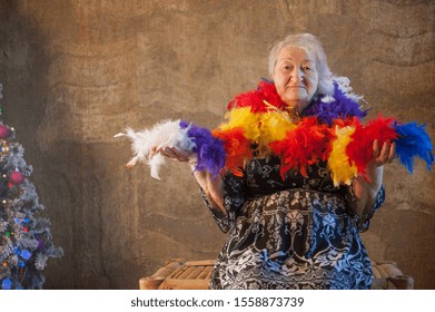 Extravagant Old Woman With Gray Hair And Wrinkles In Glasses. Senior Citizen In Carnival Hat And Colored Feathers Around Neck. Concept Of Domineering And Funny Elderly Mother In Law. Christmas Eve