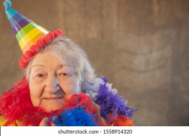 Extravagant Old Woman With Gray Hair And Wrinkles In Glasses. Senior Citizen In Carnival Hat And Colored Feathers Around Neck. Concept Of Domineering And Funny Elderly Mother In Law. Christmas Eve