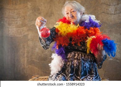 Extravagant Old Woman With Gray Hair And Wrinkles In Glasses. Senior Citizen In Carnival Hat And Colored Feathers Around Neck. Concept Of Domineering And Funny Elderly Mother In Law. Christmas Eve