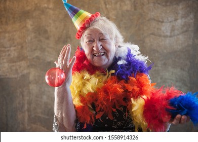 Extravagant Old Woman With Gray Hair And Wrinkles In Glasses. Senior Citizen In Carnival Hat And Colored Feathers Around Neck. Concept Of Domineering And Funny Elderly Mother In Law. Christmas Eve