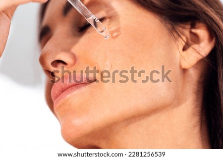 Extreme close up of beautiful smiling girl. Young woman applies transparent serum from pipette to her face. Pretty female brunette closed her eyes and relaxed during skin moisturizing procedure.