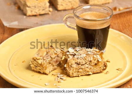 Extreme  close up of two freshly baked, homemade apricot pecan bars and a clear glass of hot espresso on a yellow, round plate and three bars, cooling on parchment paper on wood cutting board