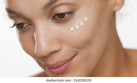 Extreme close-up beauty portrait of young gorgeous African American woman with face cream dots on her cheek under eye on white background | Face or eye cream commercial concept