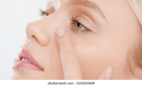 Extreme close-up of young Caucasian woman with pure skin applies face serum smearing drops on her cheek on white background | Skin care serum commercial concept