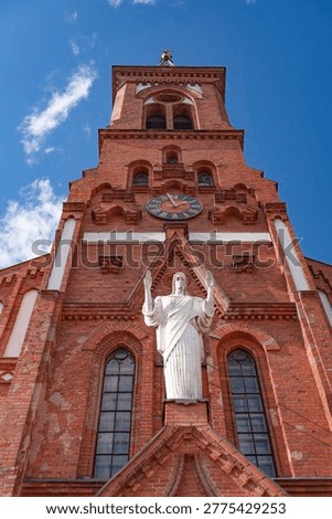 Extreme low angle shot of the neogothic Saint Anthony of Padua church in Postavy, Belarus. Red brick facade, Roman numeral clock and Jesus statue against blue sky.