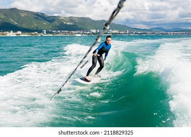 Extreme Sport On Water Concept. Young Athletic Female Wakeboarder Surfing Behind Boat. Woman In Wet Suit Practice Wakesurfing On Sea. 