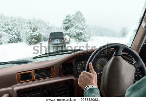 Extreme winter driving on snow off-road car travel.\
Hand of male driver on steering wheel of vehicle and view from auto\
window of suv caravans driving along wild forest road and\
snow-covered trees