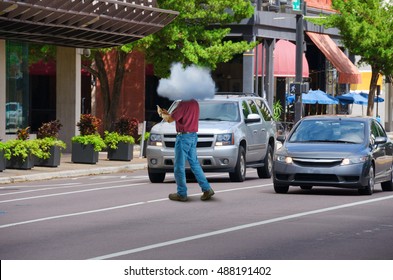 Extremely distracted cell phone using man is unknowingly walking out into city traffic jaywalking causing traffic problems; stupidity illustrated by man having his head in the clouds.