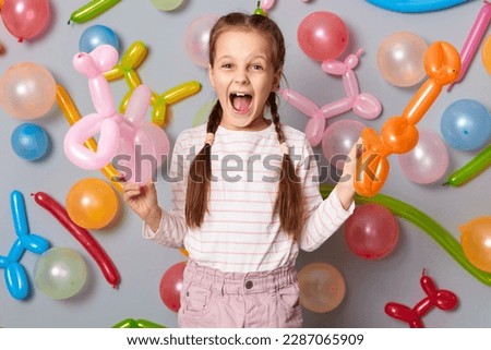 Extremely excited festive little girl  with pigtails standing against gray decorated colorful wall holding balloons in hands screaming hurray celebrating birthday.