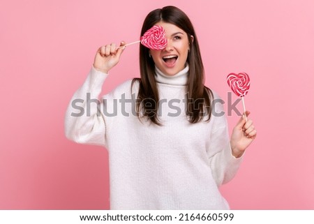 Extremely happy playful brunette woman covering eye with delicious candy in heart shape, looks away, wearing white casual style sweater. Indoor studio shot isolated on pink background.