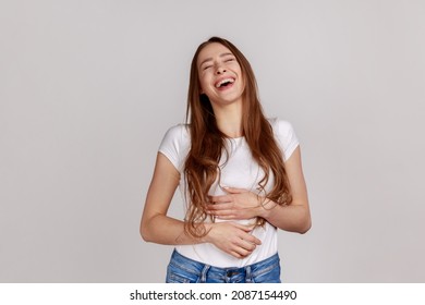 Extremely happy woman holding her stomach and laughing out loud, chuckling giggling at amusing anecdote, sincere emotion, wearing white T-shirt. Indoor studio shot isolated on gray background.