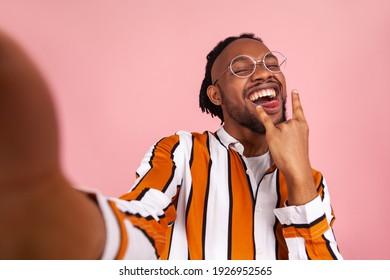 Extremely Satisfied African Man With Dreadlocks In Striped Shirt Making Tongue Out Showing Rock And Roll Gesture, Posing At Selfie Camera, Having Fun. Indoor Studio Shot Isolated On Pink Background