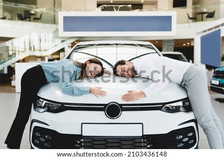Extremely satisfied happy caucasian young couple family husband and wife hugging embracing their new car, feeling excited after buying expensive auto at auto dealer shop store.