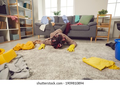 Extremely tired woman lying face down on rug in living-room amid chaos of scattered untidy clothes. Miserable exhausted housewife fallen on floor while cleaning and tidying crazy mess in her house