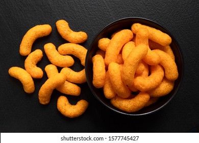 Extruded cheese puffs in a black ceramic bowl next to spilled cheese puffs isolated on black. Top view.