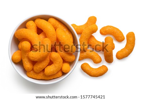 Extruded cheese puffs in a white ceramic bowl next to spilled cheese puffs isolated on white. Top view.
