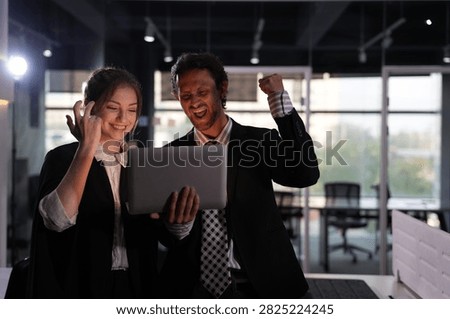 Exuberant cries echo through the empty workspace while the bearded man pumps his fist and the young businesswoman flings her fingers wide over the laptop in a bright modern coworking office.