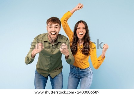 Exuberant man and woman cheerfully celebrating a victory, punching the air with raised fists, against a vibrant blue backdrop, looking at camera