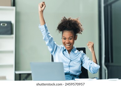 Exuberant and triumphant, a young African American woman in a blue formal shirt with afro brown hair operates a tablet, notebook, and mobile phone in a modern office setting. - Powered by Shutterstock