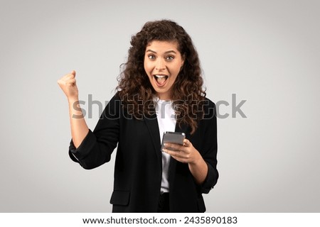 Exuberant young businesswoman with curly hair holding smartphone and making triumphant fist pump, expressing jubilation or success, against grey backdrop