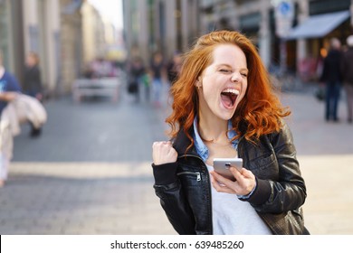 Exuberant young woman cheering at good news on her mobile phone and punching the air with her fist on an urban street with copy space