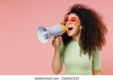 Exultant happy vivid young curly latin woman 20s wears mint t-shirt sunglasses hold scream in megaphone announces discounts sale Hurry up isolated on plain pastel light pink background studio portrait