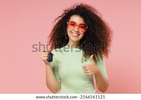 Exultant happy young curly latin woman 20s wears mint t-shirt sunglasses hold vehicle key show thumb up like gesture isolated on plain pastel light pink background studio portrait. Car sales concept