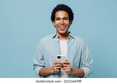 Exultant Young Black Curly Man 20s Years Old Wears White Shirt Hold In Hand Use Mobile Cell Phone Typing Browsing Chatting Looking Camera Isolated On Plain Pastel Light Blue Background Studio Portrait