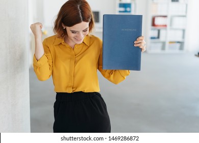Exultant young woman celebrating getting a job after a successful interview, smiling and punching the air with her fists while holding her CV. Bewerbung is german word for application file