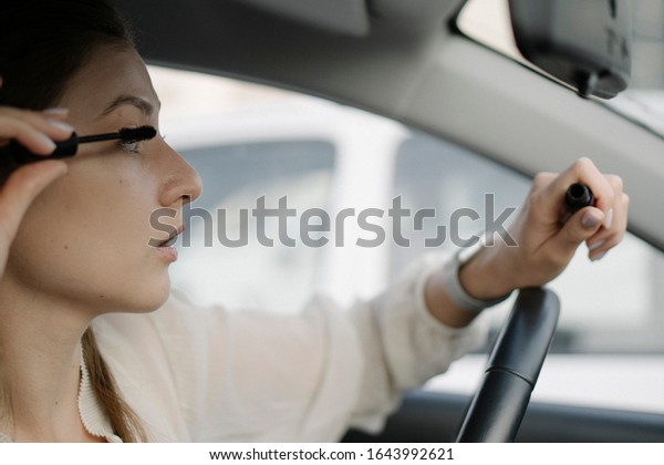 Eye brush, shadows, make-up in the car. A young\
woman looks in the mirror inside the car and applies make-up before\
a business meeting