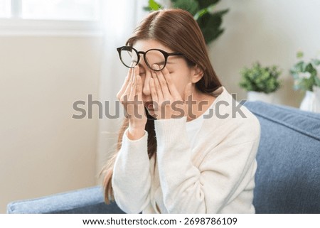 Eye care problem concept, fatigue asian young woman tired eyes irritated rubbing, massage eye dry relief vision pain sitting on couch, sofa at home, eye strain, hand holding glasses, suffer bad blurry