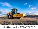 Eye catching yellow road roller with enclosed climate controlled cabin stands on not ready new road, stones, blue sky, clouds, left side view. Clean shiny old heavy tractor
