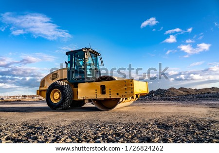 Eye catching yellow road roller with enclosed climate controlled cabin stands on not ready new road, stones, blue sky, clouds, left side view. Clean shiny old heavy tractor