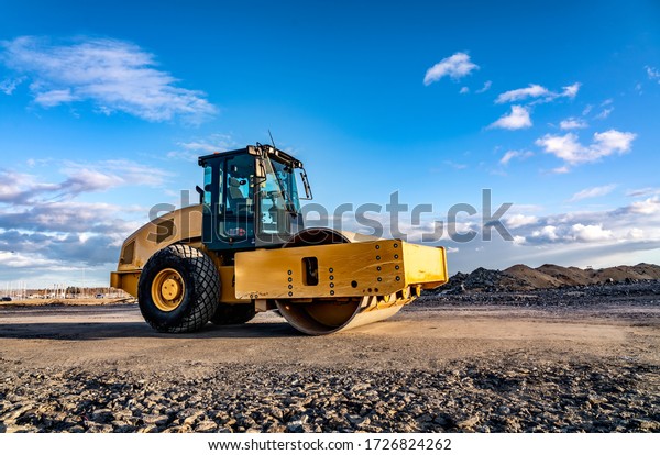 Eye catching yellow\
road roller with enclosed climate controlled cabin stands on not\
ready new road, stones, blue sky, clouds, left side view. Clean\
shiny old heavy tractor