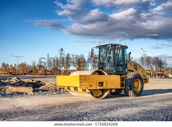 Eye catching\
yellow road roller with enclosed climate controlled cabin stands on\
not ready new road, stones, blue sky, clouds, front right side\
view. Clean shiny old heavy\
tractor