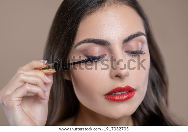 Eye\
cosmetics. Face of serious involved beautiful woman with drooping\
eyelids holding brush with mascara near her\
eye