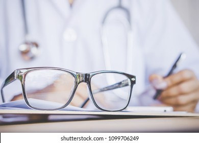 Eye Glasses On Desk With Blur Background Of Doctor Writing Prescription ,working In Hospital Office 