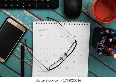 Eye glasses on desktop calendar surrounded by personal computer, blank screen smartphone, stationary, and a glass of water. Top view of messy work desk
