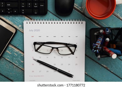 Eye Glasses And Pen On Desktop Calendar, Surrounded By Personal Computer, Blank Screen Smartphone, Stationary, And Glass Of Water. Top View Of Messy Work Desk