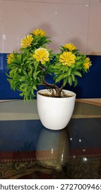 An eye level medium shot captures a small, decorative artificial plant with bright yellow flowers and lush green fern like leaves, housed in a simple, glossy white ceramic pot. The plant is situated on a reflective glass tabletop, which shows a clear reflection of the pot and base. The background consists of a wall with blue and white tiles, providing a clean, modern contrast. The image highlights the use of artificial greenery and floral for indoor settings, offices, or home staging. It represents themes of interior design, decoration, maintenance free plants, and modern home.