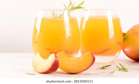An eye level shot features two stemless glasses filled with vibrant yellow peach juice, decorated with a sprig of fresh rosemary. Slices of juicy peach lie scattered on a bright marble surface.
