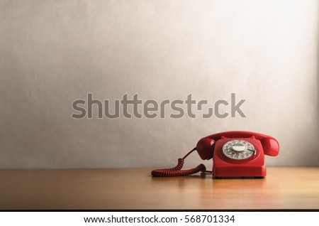Eye level shot of a retro red telephone (British circa 1960s to 1970s) on a light wood veneer desk or table with off white background providing copy space.