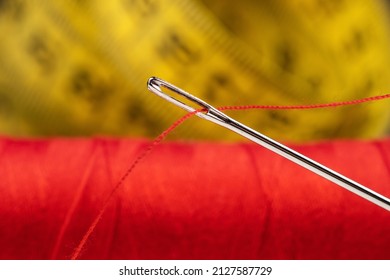 Eye Of The Needle With Red Polyester Thread. Spool Of Red Polyester Sewing Thread. Measuring Tape In The Background.