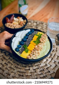 Bird’s Eye View Of A Green Vegan Smoothie Bowl With Tropical Fruits  Granola Like Mango, Spirulina,  Coconut, Nuts,  Chia Seeds, Mint On A Wooden Table Eating A Healthy Breakfast With A Spoon