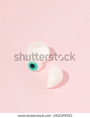 Eyeball falling out of cracked eggshell, minimal, creative Halloween celebration layout on pastel pink background.