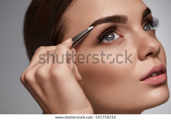 Eyebrow Correction. Closeup Of Beautiful Young\
Woman With Perfect Makeup And Long Lashes Plucking Eyebrows.\
Portrait Of Sexy Female Model Face And Tweezers Near Brows. Beauty\
Concept. High\
Resolution