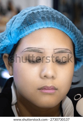 Eyebrow tattooing process shown on young woman with closed eyes wearing blue hair cap in beauty salon, preparation for semi permanent makeup treatment