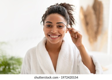 Eyebrows Plucking. Happy African American Woman Tweezing And Styling Her Brows With Tweezers Smiling Looking At Camera Posing Standing In Modern Bathroom At Home. Female Beauty Routine