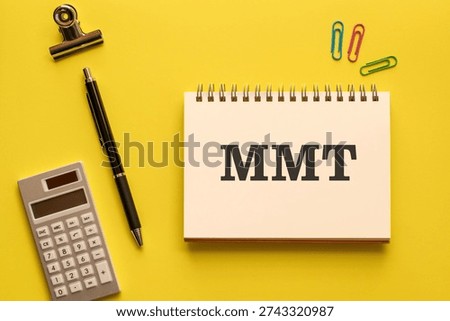 Eye-catching image of MMT text on notebook. The word stands for Modern Monetary Theory. Shown with calculator, pen, clips.